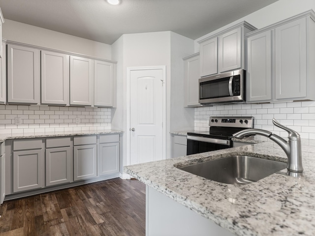 kitchen with a sink, appliances with stainless steel finishes, light stone countertops, dark wood-type flooring, and gray cabinets