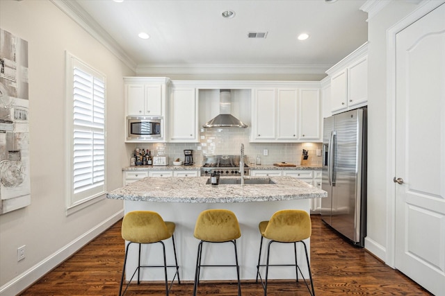 kitchen with white cabinets, appliances with stainless steel finishes, a sink, an island with sink, and wall chimney exhaust hood