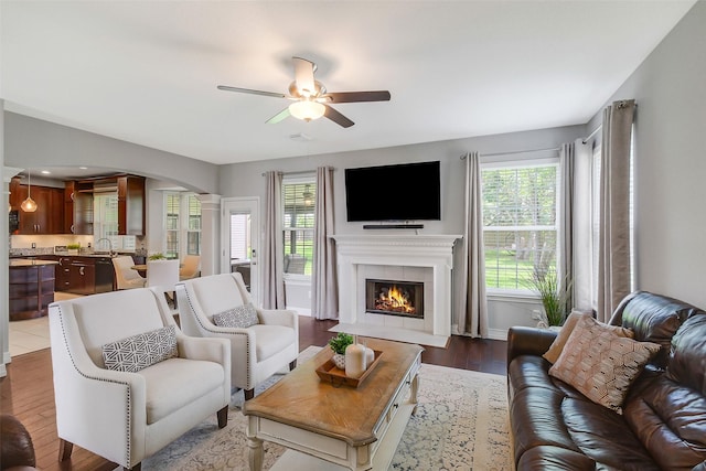 living room featuring a tile fireplace, a ceiling fan, ornate columns, arched walkways, and wood finished floors