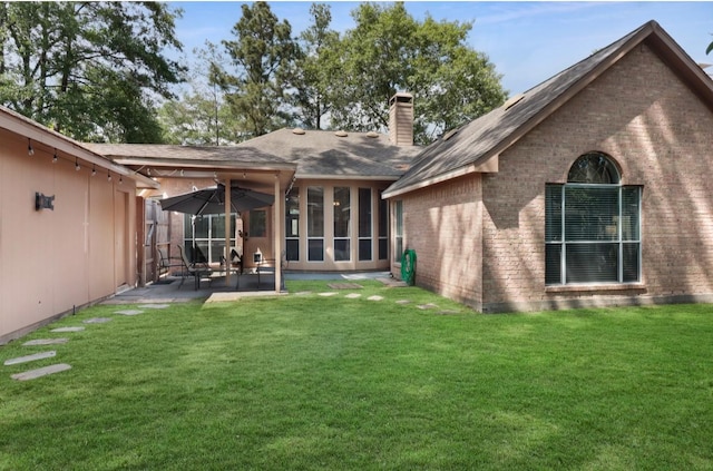 rear view of property featuring a patio area, a yard, a chimney, brick siding, and roof with shingles