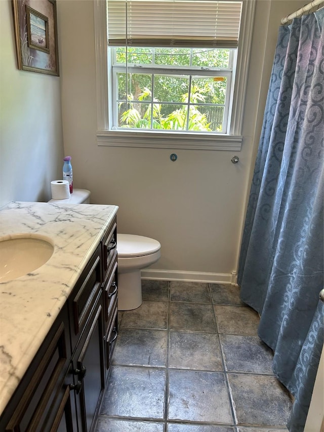 bathroom featuring stone tile flooring, a shower with shower curtain, toilet, vanity, and baseboards