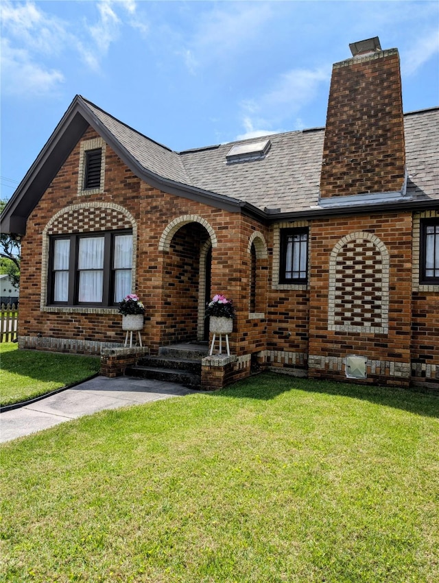 tudor-style house with brick siding, a shingled roof, a front lawn, and a chimney