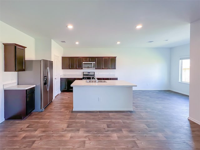 kitchen featuring wood finished floors, dark brown cabinetry, an island with sink, a sink, and appliances with stainless steel finishes