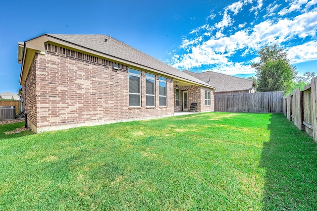 rear view of house featuring a fenced backyard, roof with shingles, brick siding, a lawn, and central air condition unit