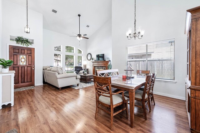 dining room with visible vents, wood finished floors, high vaulted ceiling, a tile fireplace, and ceiling fan with notable chandelier