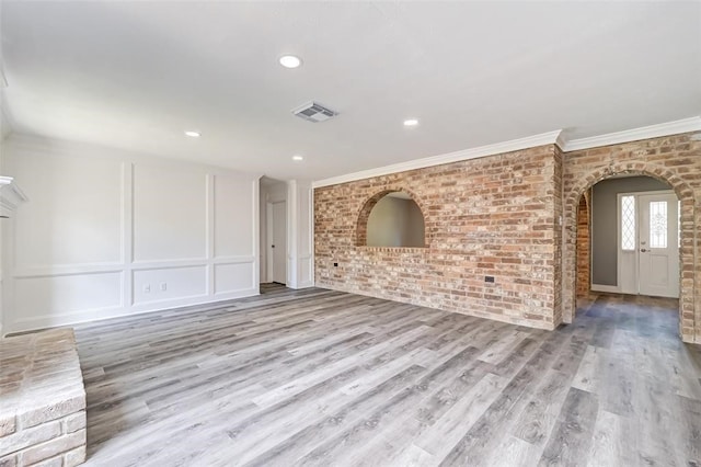 unfurnished living room featuring arched walkways, brick wall, wood finished floors, visible vents, and ornamental molding
