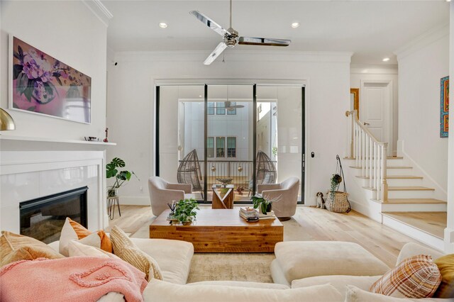 living room with crown molding, stairs, a glass covered fireplace, wood finished floors, and a ceiling fan