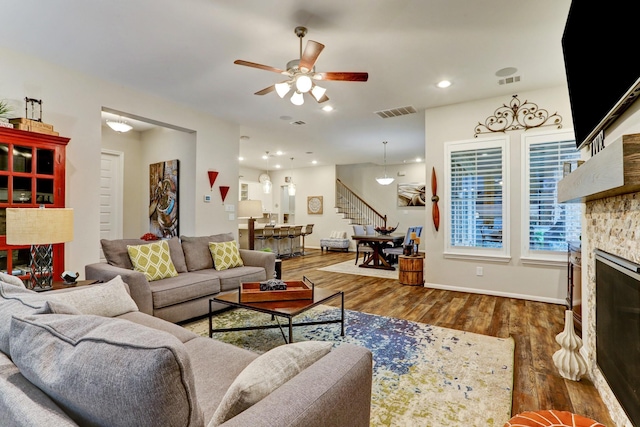 living room with ceiling fan, a stone fireplace, visible vents, wood finished floors, and stairway