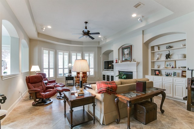 living room with visible vents, a fireplace, a raised ceiling, built in shelves, and a ceiling fan