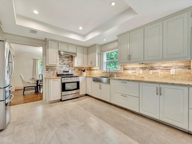 kitchen with appliances with stainless steel finishes, a sink, under cabinet range hood, a tray ceiling, and backsplash