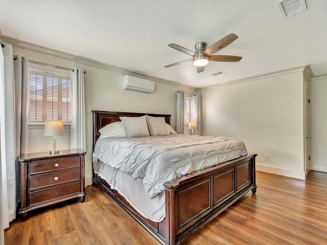 bedroom featuring a wall unit AC, light wood finished floors, crown molding, baseboards, and a ceiling fan