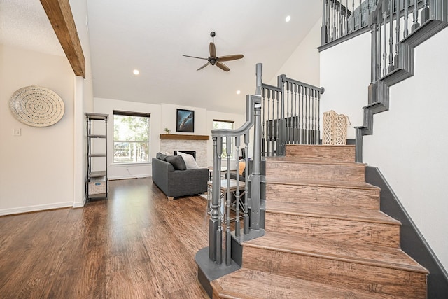 staircase featuring ceiling fan, a fireplace, wood finished floors, baseboards, and high vaulted ceiling