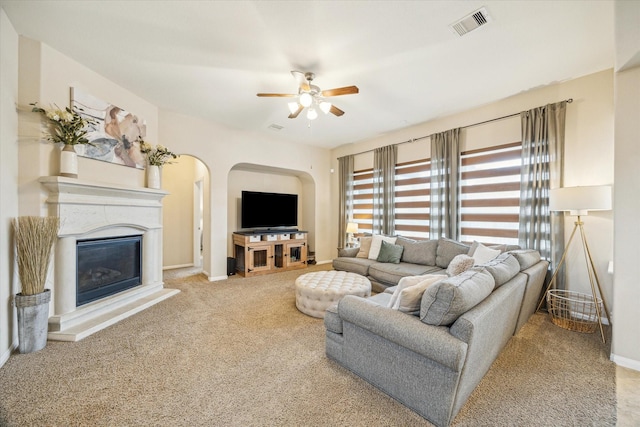 carpeted living area featuring ceiling fan, arched walkways, a glass covered fireplace, baseboards, and visible vents
