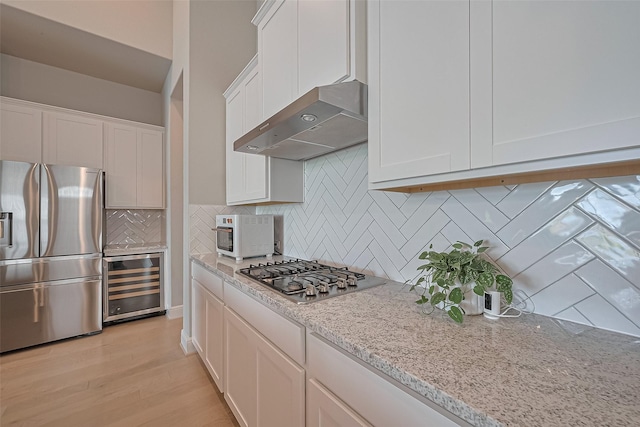 kitchen featuring wine cooler, wall chimney range hood, white cabinets, light stone counters, and stainless steel appliances