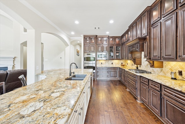 kitchen with light stone countertops, appliances with stainless steel finishes, a sink, arched walkways, and glass insert cabinets