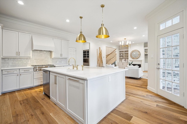 kitchen featuring stainless steel appliances, a kitchen island with sink, light wood-style floors, premium range hood, and tasteful backsplash