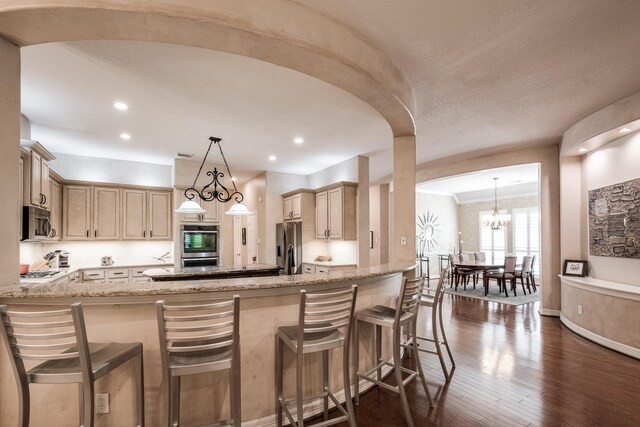 kitchen with dark wood-style flooring, light stone counters, an inviting chandelier, stainless steel appliances, and a peninsula