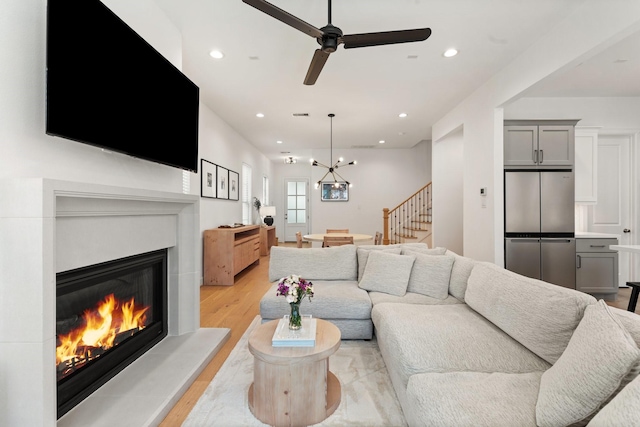 living area with a glass covered fireplace, light wood-style flooring, ceiling fan with notable chandelier, recessed lighting, and stairway