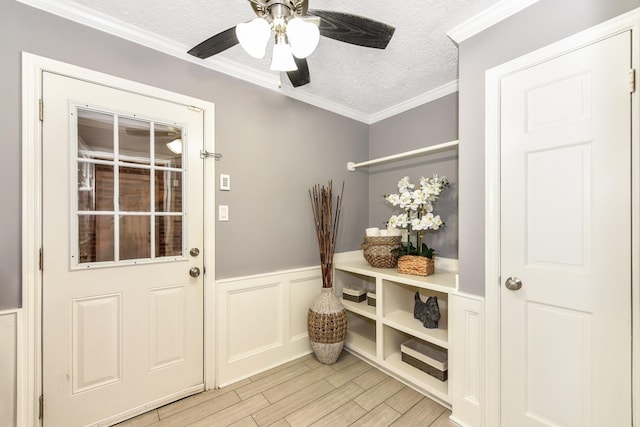 doorway with ceiling fan, a textured ceiling, crown molding, a wainscoted wall, and light wood-style floors