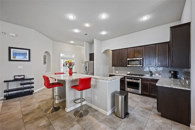 10418 Texas Sage Way, Cypress, TX 77433 - kitchen with appliances with stainless steel finishes, arched walkways, decorative backsplash, dark brown cabinetry, and a kitchen island with sink