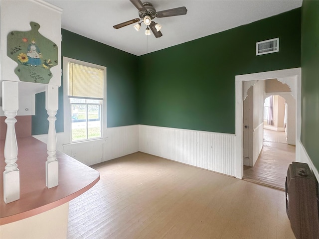 unfurnished room featuring ceiling fan, visible vents, arched walkways, wood finished floors, and a wainscoted wall