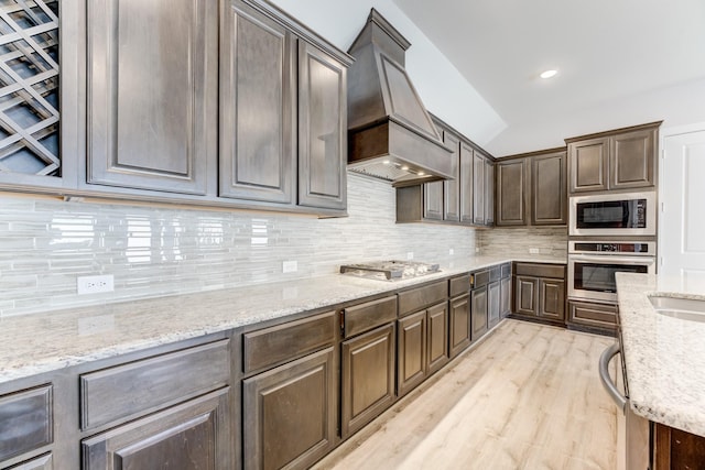 kitchen featuring decorative backsplash, dark brown cabinetry, appliances with stainless steel finishes, light wood-style floors, and custom range hood