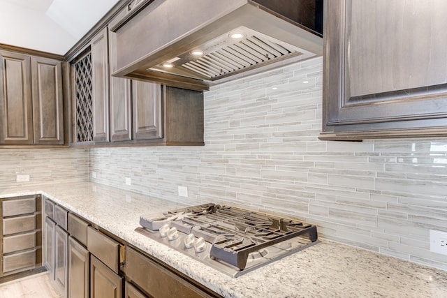kitchen with decorative backsplash, stainless steel gas cooktop, custom exhaust hood, dark brown cabinetry, and light stone countertops
