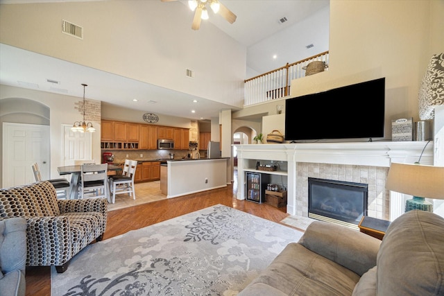 living room featuring arched walkways, a tiled fireplace, ceiling fan with notable chandelier, light wood-type flooring, and visible vents