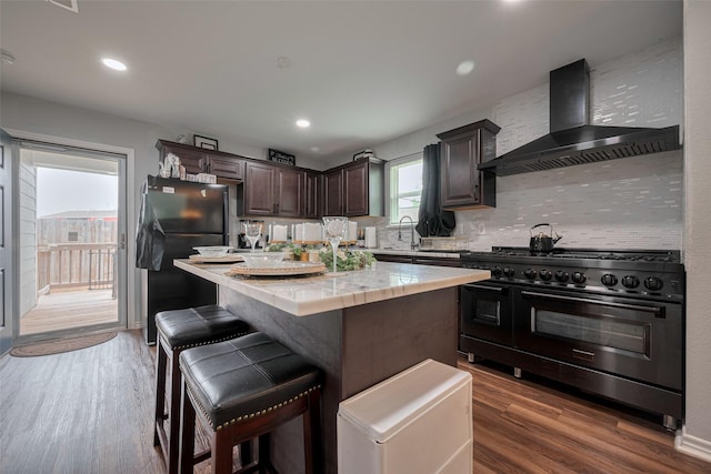 kitchen featuring wall chimney range hood, backsplash, dark brown cabinets, light countertops, and black appliances