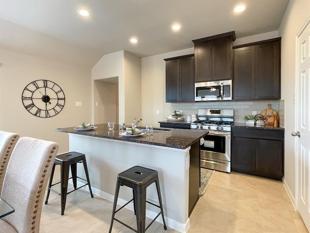 kitchen featuring a sink, an island with sink, dark stone counters, stainless steel appliances, and tasteful backsplash