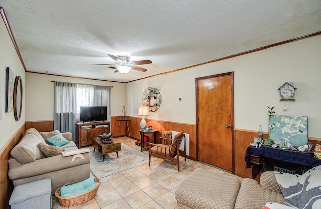 living room featuring crown molding, wood walls, a textured ceiling, a wainscoted wall, and a ceiling fan