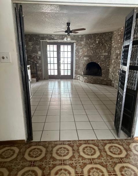 unfurnished living room with french doors, a ceiling fan, a textured ceiling, a stone fireplace, and light tile patterned flooring