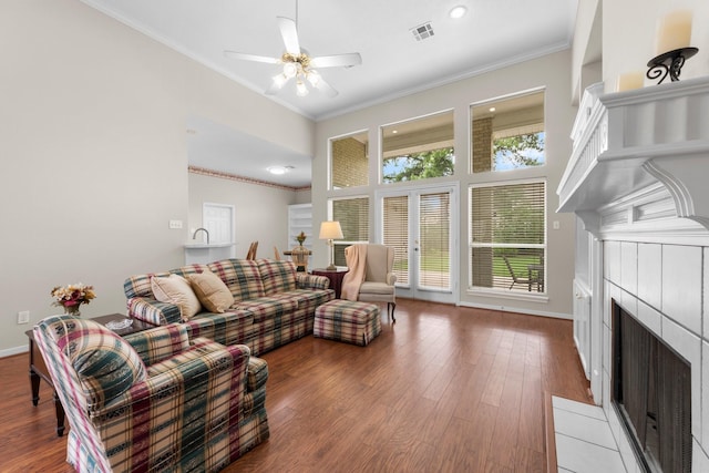 living area featuring ceiling fan, a tiled fireplace, wood finished floors, visible vents, and ornamental molding