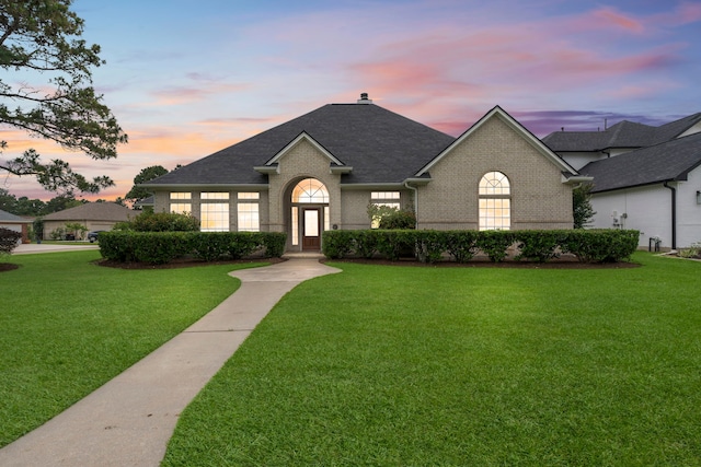 french provincial home featuring roof with shingles, a chimney, a lawn, and brick siding