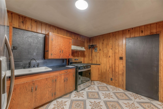 kitchen featuring brown cabinetry, a sink, stainless steel gas range, wooden walls, and under cabinet range hood