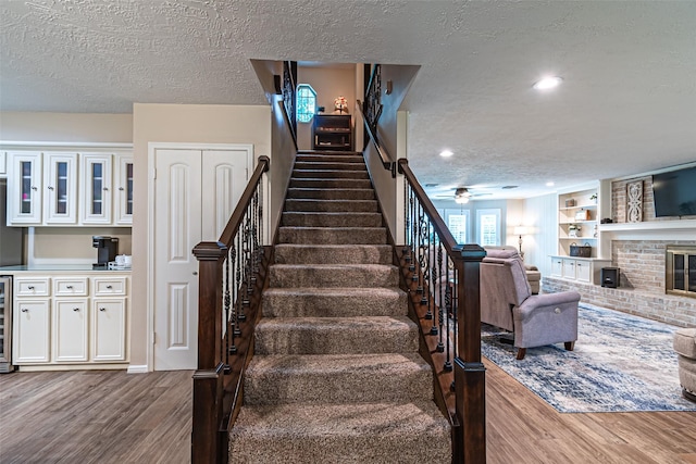 stairs with ceiling fan, a brick fireplace, beverage cooler, a textured ceiling, and wood finished floors