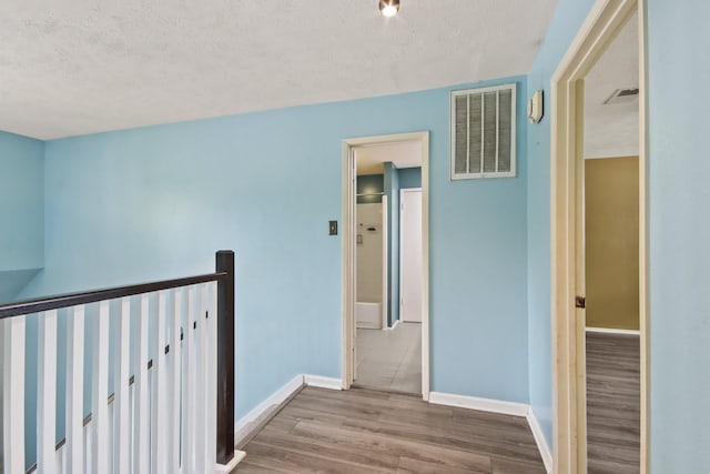 corridor with a textured ceiling, visible vents, wood finished floors, baseboards, and an upstairs landing