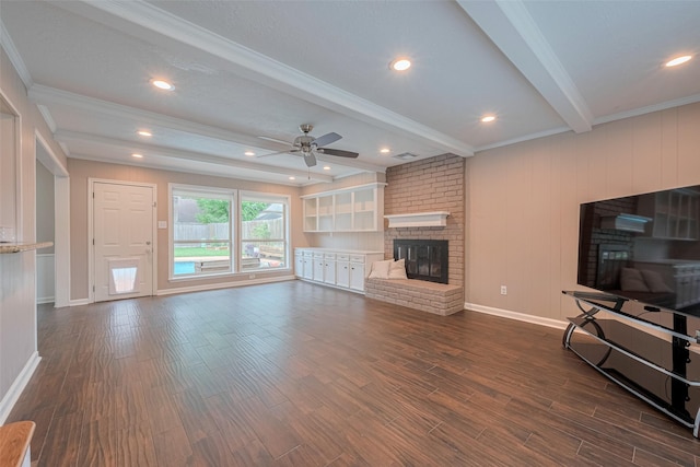 unfurnished living room featuring a ceiling fan, crown molding, a brick fireplace, beam ceiling, and dark wood-type flooring