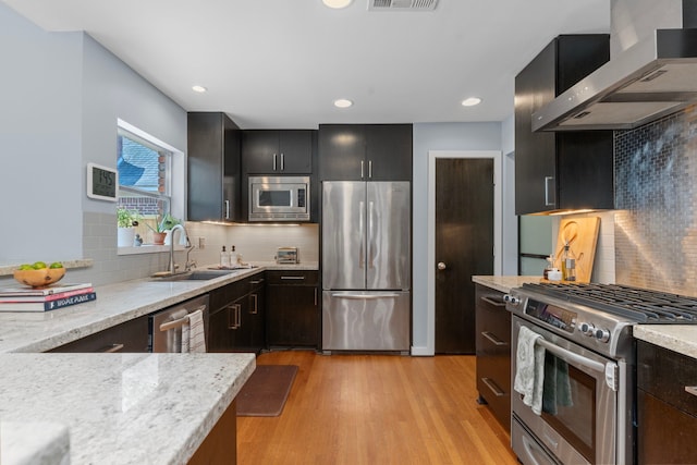 kitchen featuring wall chimney range hood, appliances with stainless steel finishes, a sink, light wood-type flooring, and backsplash