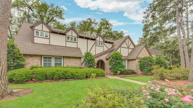 tudor-style house featuring a shingled roof, stucco siding, a front lawn, and brick siding