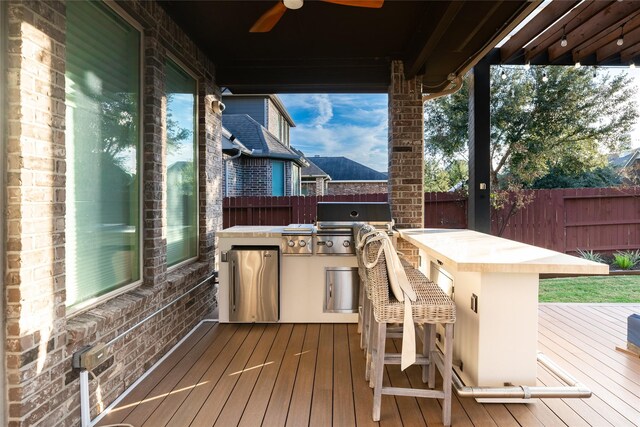wooden terrace featuring area for grilling, ceiling fan, fence, outdoor wet bar, and an outdoor kitchen