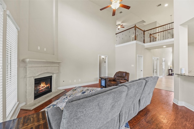 living room with ceiling fan, a towering ceiling, wood finished floors, baseboards, and a glass covered fireplace