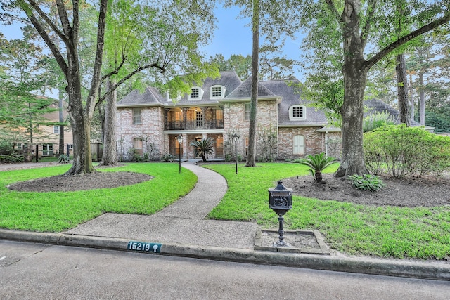 french country style house with brick siding, stone siding, a front lawn, and a balcony
