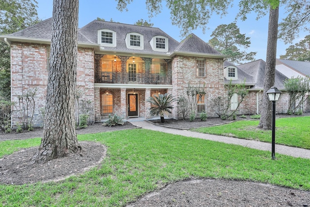 french provincial home featuring brick siding, a balcony, a front yard, and roof with shingles