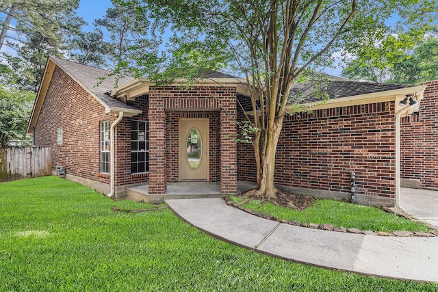 view of exterior entry with brick siding, roof with shingles, fence, and a yard