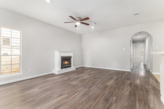 unfurnished living room featuring a ceiling fan, a lit fireplace, visible vents, arched walkways, and dark wood-style floors