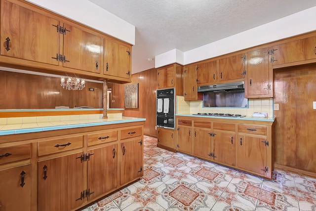 kitchen featuring brown cabinetry, backsplash, under cabinet range hood, stainless steel gas stovetop, and black oven