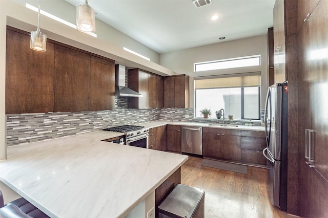 kitchen featuring a peninsula, wood finished floors, wall chimney range hood, a sink, and appliances with stainless steel finishes
