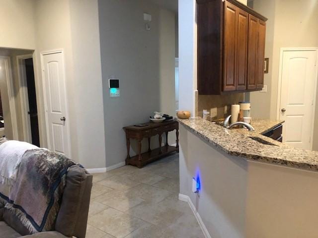 kitchen with brown cabinetry, a peninsula, light stone countertops, baseboards, and light tile patterned floors