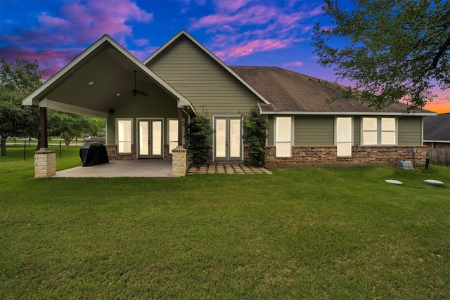 back of house at dusk with brick siding, a patio area, french doors, a ceiling fan, and a lawn